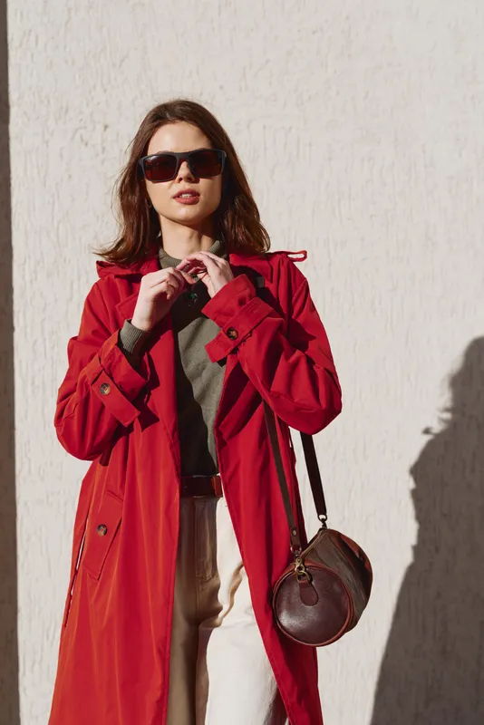 A stylish young woman in a red trench coat stands outdoors, adjusting her collar while wearing dark sunglasses, with a pale wall and strong sunlight behind her.