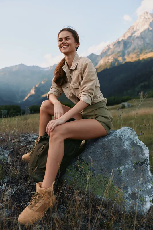 Cheerful woman outdoors, smiling on a rock with sunlit mountains in the background, casual outfit, boots and shorts, natural colors, gentle breeze, open landscape