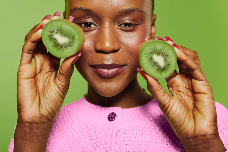 Close-up of smiling dark skinned African woman holding two halves of fresh kiwi fruit near face. Concept of healthy lifestyle, fresh food, natural vitamins, and wellness on green background.