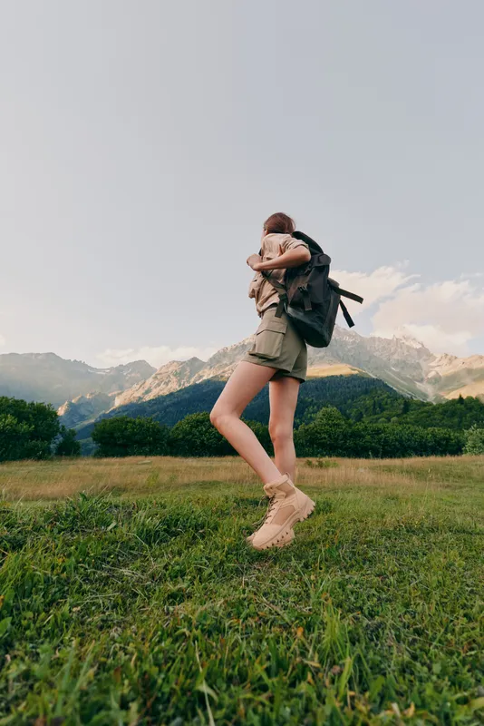 Cheerful woman outdoors, mid jump with backpack, hiking style in a green meadow, mountains in the background, warm sunlight, energetic lifestyle moment, natural colors