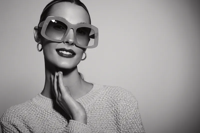 Stylish smiling caucasian woman with fair skin wearing large sunglasses and hoop earrings poses with hand on neck against plain background. Fashion and beauty concept.