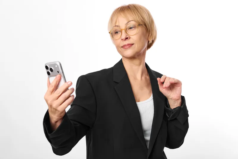 Senior woman with glasses in black blazer holds smartphone, gestures toward camera, clean white background, concept for modern business technology and communication