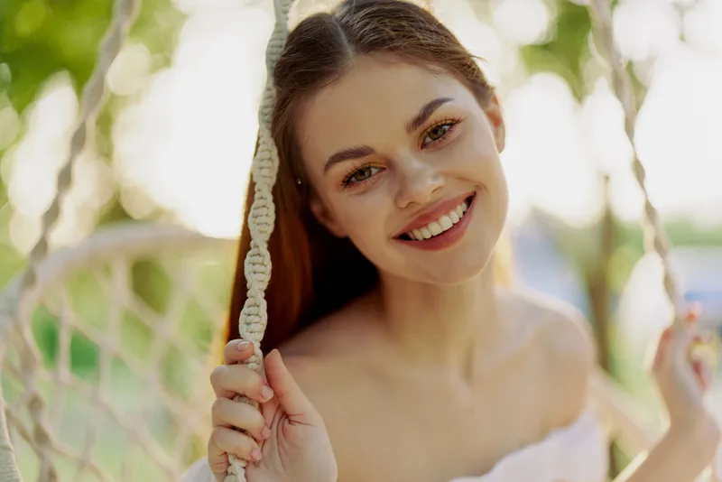 Young woman with a joyful smile posing on a swing, wearing light summer attire in a vibrant outdoor setting, captured in warm natural light