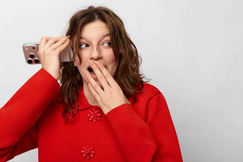 Surprised young woman in red sweater holds smartphone near ear, covering mouth with hand. Isolated on white background. Concept for unexpected news, shock, and communication.