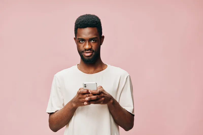 Young Black man in a light t shirt engaging with a smartphone against a soft pink background, showcasing modern technology and social interaction