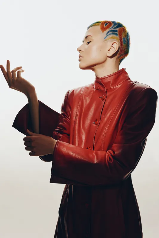 Stylish woman with colorful short hair wearing a red leather jacket poses thoughtfully against a plain light background, showcasing modern fashion and bold hairstyle.
