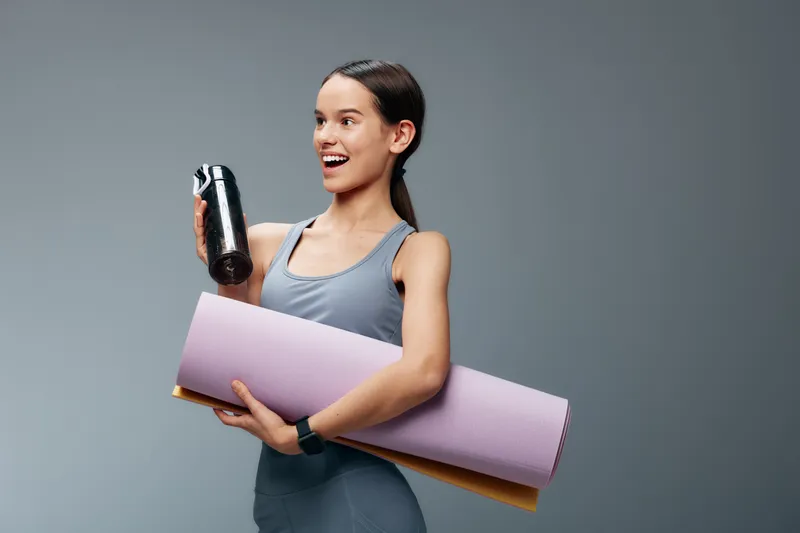 Fitness minded woman in athletic wear holds a water bottle and yoga mat, ready for a workout. Energetic studio vibe emphasizing exercise, training, and healthy lifestyle.