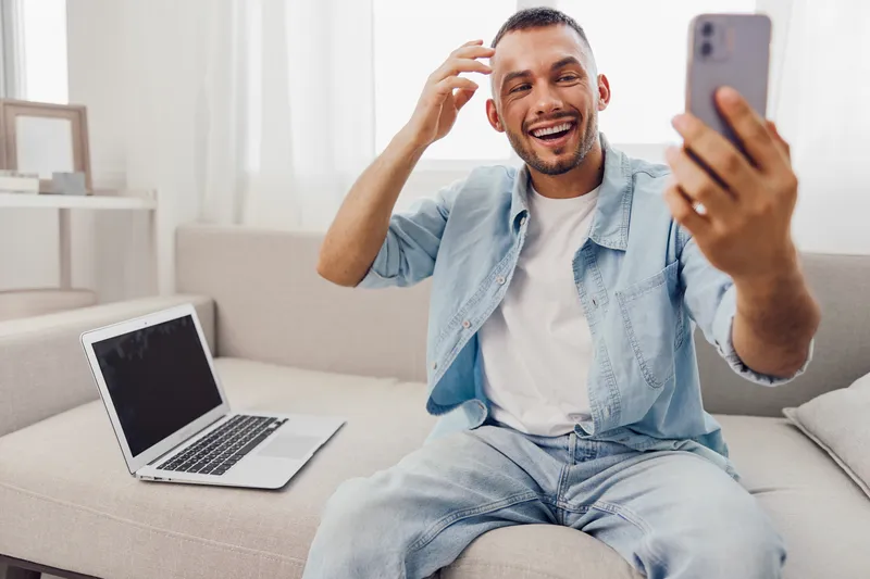 Joyful man engaging with technology, blending work and personal life while smiling at his smartphone in a cozy home environment, promoting mindfulness.