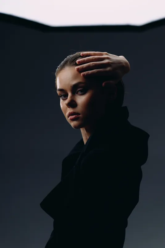 Moody woman in black coat with hand on head against dark background, dramatic lighting