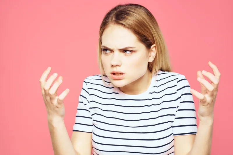Блондинка в полосатой футболке выражает недовольство с помощью жестов и мимики на розовом фоне