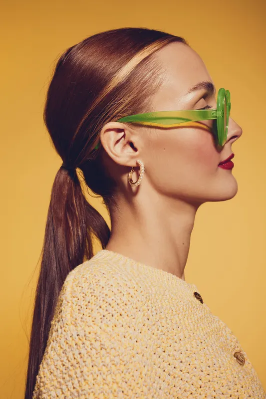 Profile of a stylish woman with long brown hair tied back wearing green sunglasses and gold hoop earrings against a warm yellow background. Fashion and summer concept.