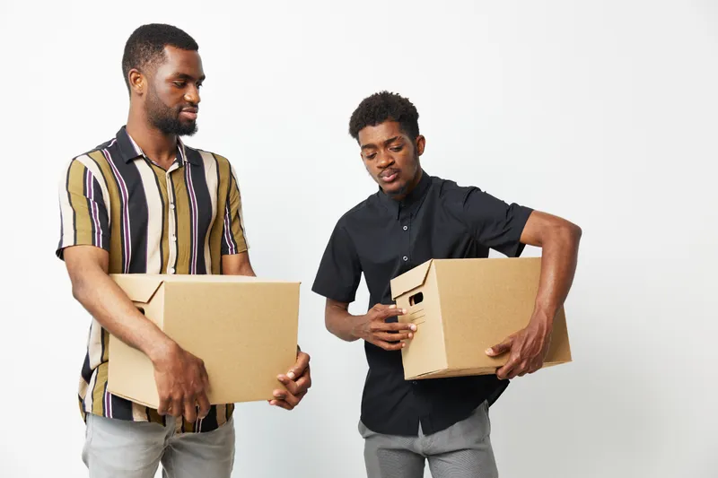 Two African American men are collaborating while carrying cardboard boxes, showcasing teamwork and productivity in a modern workspace. Bright, engaging atmosphere.