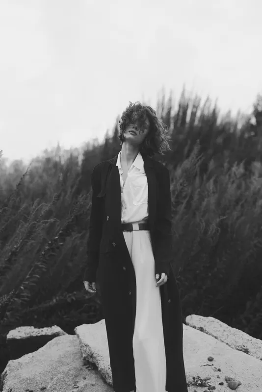 Monochrome scene of a stylish woman in a long coat standing on rocky terrain beside tall grasses, conveying fashion, confidence and urban elegance in a natural setting