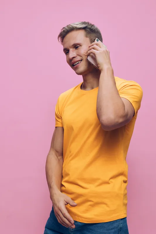Smiling man having a phone conversation against a pink background, dressed in a bright yellow t-shirt and blue jeans, casual style, happy mood, lifestyle shot, colorful clothing, expression of joy and