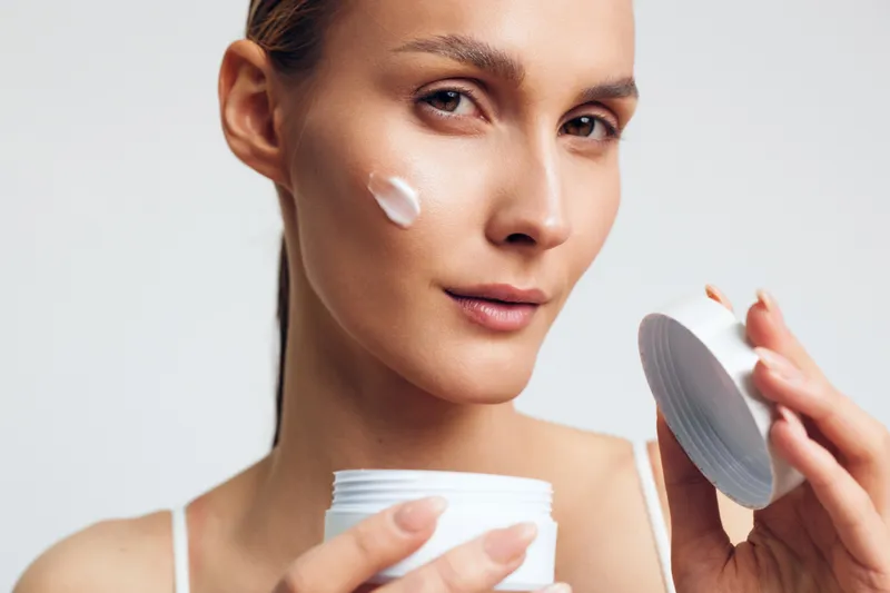 Woman applying cream to her face, moisturizer jar in hand, clean studio portrait with soft lighting, calm expression, promoting skincare routine and beauty care