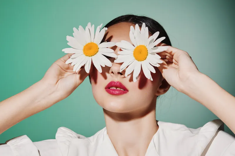 Young woman with red lipstick hides eyes behind two large daisies on a green background wearing white blouse. Creative beauty and fashion concept with floral accessory and bright colors.