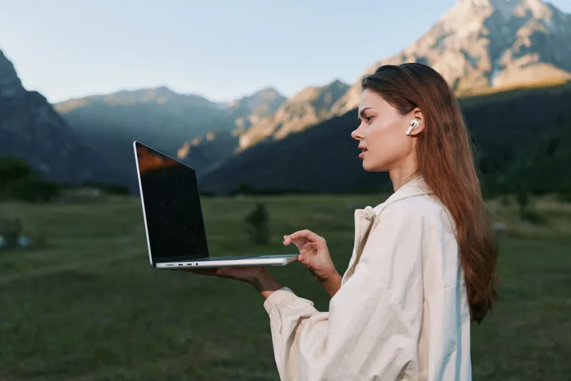 A focused professional woman stands outdoors in a scenic mountainous landscape, using a laptop. She exhibits concentration and confidence, blending technology with nature.
