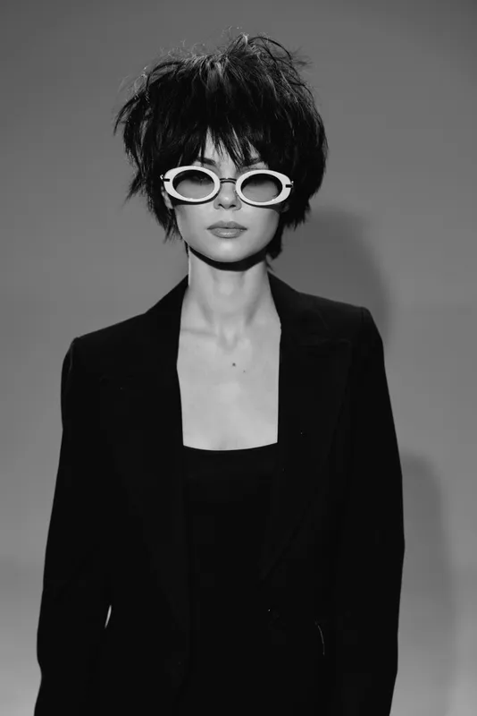 Stylish woman wearing black suit and white sunglasses with short messy hair in black and white portrait.