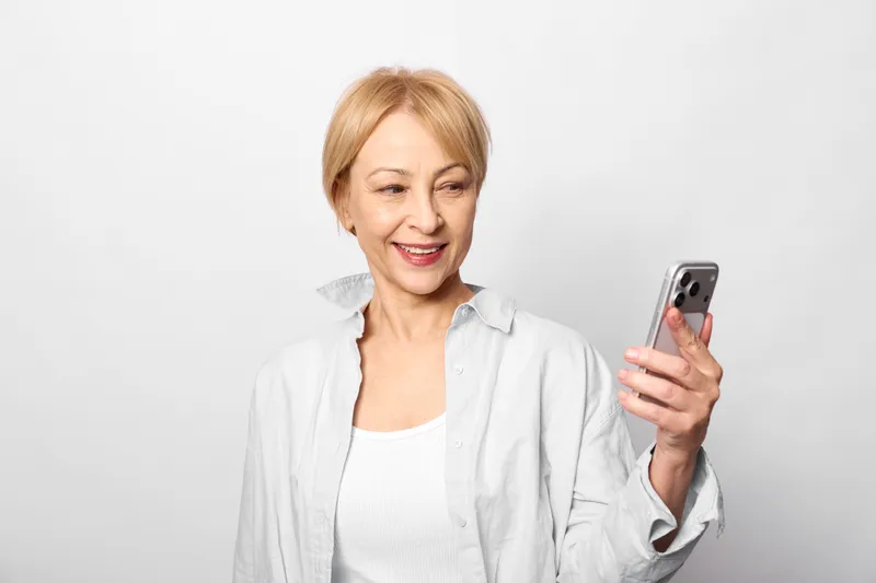 Woman elderly with smartphone smiling, portrait on gray background, holding mobile device for communication and technology, casual blouse, relaxed indoor.