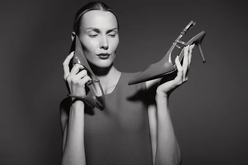 Caucasian woman with light skin holds two high heel shoes near face in black and white studio. Concept of fashion, elegance, footwear, and style.