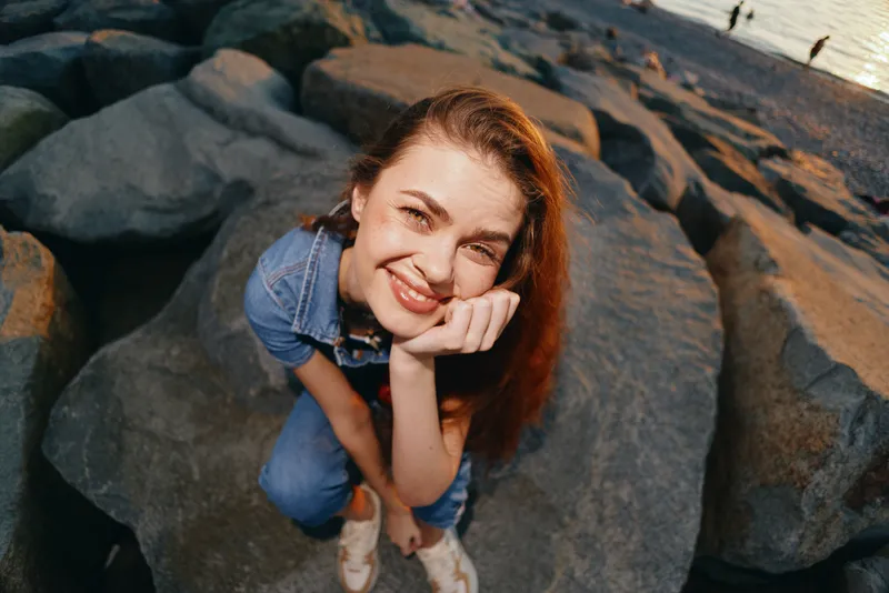lifestyle woman in modern boho-western denim street style sitting on rocks by water, smiling happily, captured with warm film color tones for a natural feel