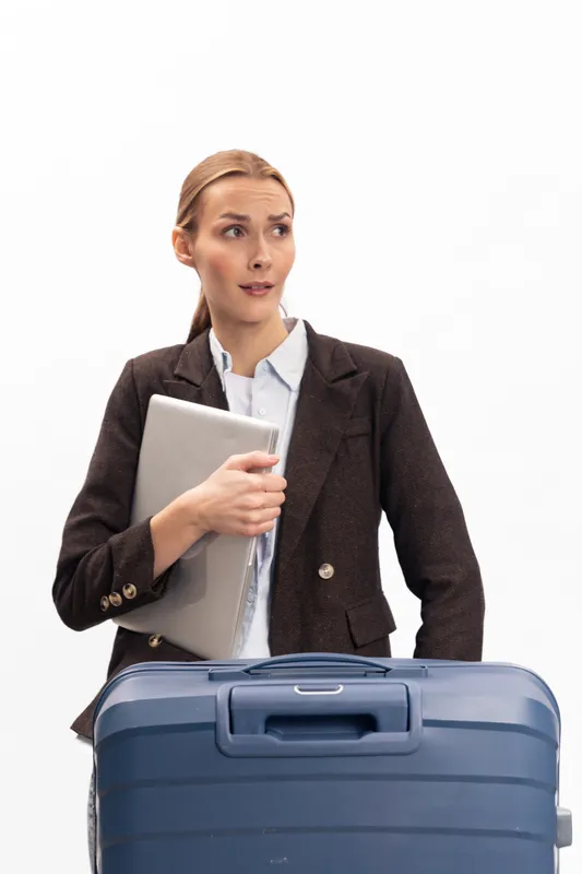 Businesswoman in formal suit holds laptop and looks aside with uncertain expression standing behind large blue suitcase on white background. Concept of travel, business trip, and decision making.