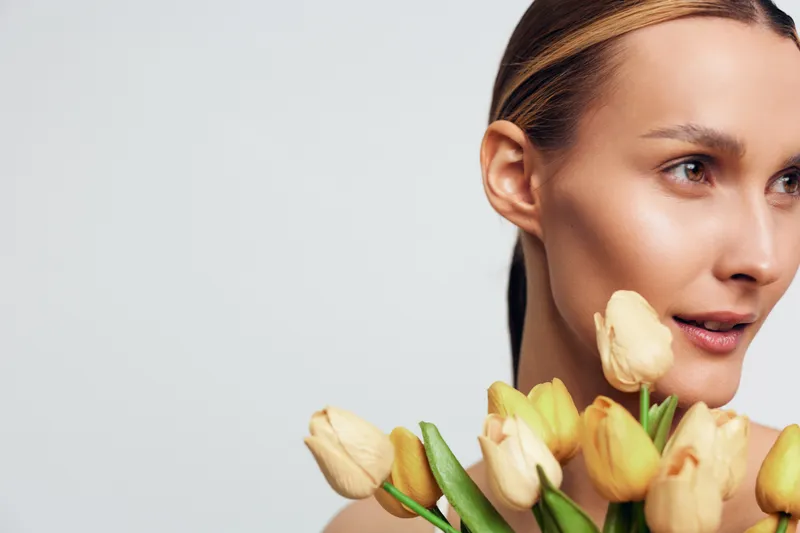 A young woman with clear skin holds a bouquet of yellow tulips close to her face, looking thoughtfully to the side against a plain light background.