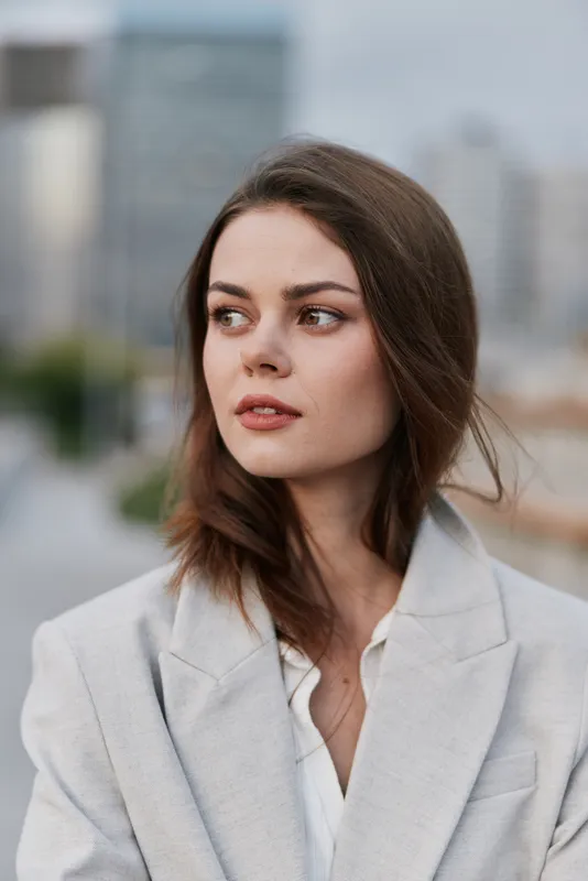 Woman in light gray blazer looking away thoughtfully with urban cityscape background, soft natural colors, modern business style, emotional expression
