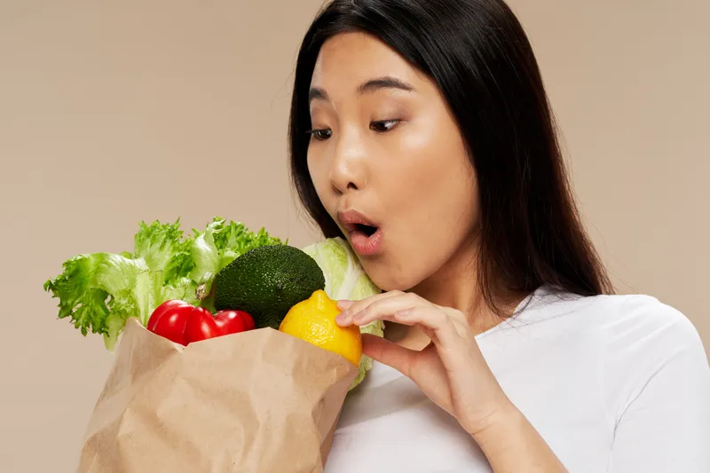 Удивлённая азиатская женщина смотрит на упаковку продуктов, рядом с ней — фигура, символизирующая диету и подсчёт калорий