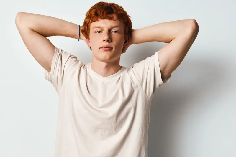 Young man with red hair wearing light t shirt standing relaxed with hands behind head on white background. Casual portrait of confident male model with neutral expression indoors.