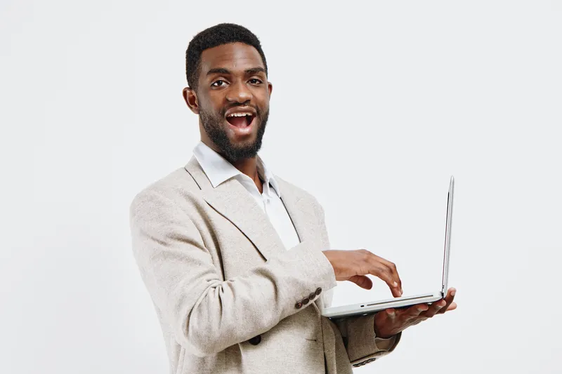Smiling young man with African descent holding a laptop computer in hand, wearing a beige blazer and white shirt, looking at camera, joyful expression, isolated on plain white background, studio portr