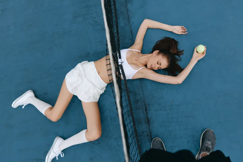 Young female tennis player in a white outfit lying on the court, expressing exhaustion after a game, with a tennis ball in hand, showcasing a vibrant athletic energy