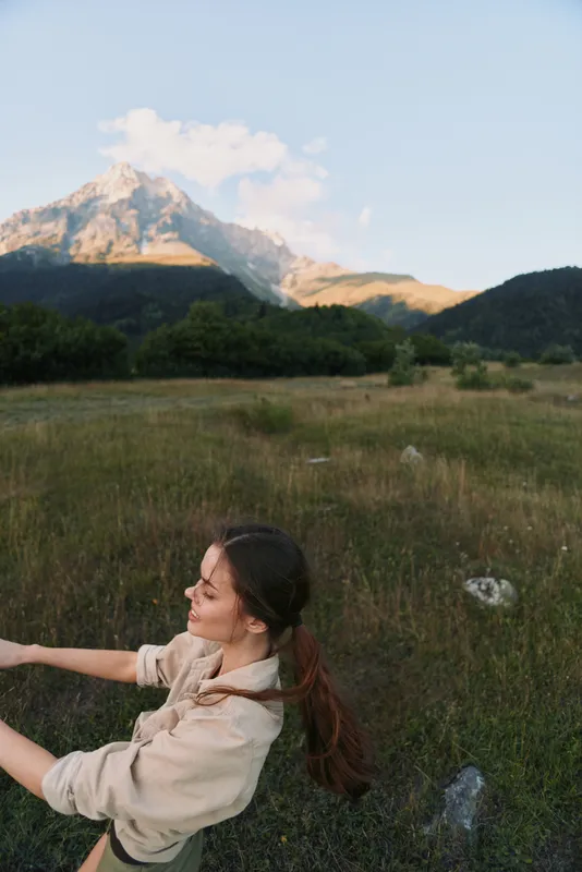 Young woman in a beige hoodie sits in a meadow with distant mountains, reaching forward with a gentle smile as warm sun lights the landscape.