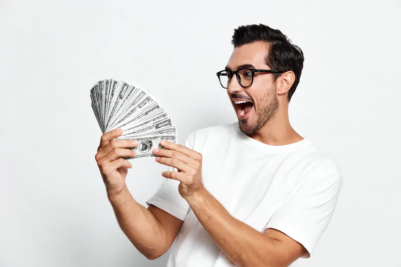 Excited young smiling man in white t-shirt, wearing glasses, holds fan of cash with joyful expression, isolated on light gray background. Lifestyle and success concept.