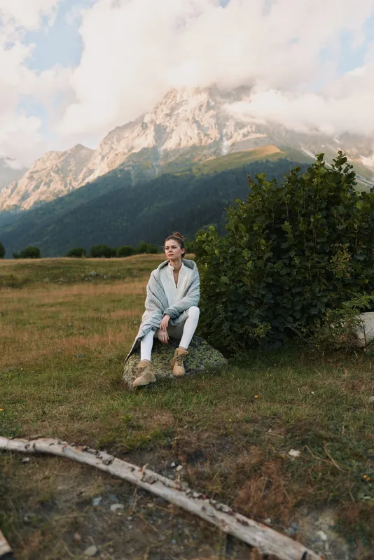 Woman sitting outdoors in a mountain landscape, lifestyle portrait with calm mood, wearing a light jacket and boots, amid natural scenery and clear sky