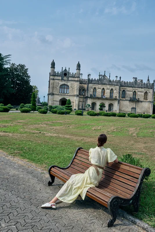 Woman in a light dress sits on a wooden bench in a park, with a historic castle in the background, creating a peaceful outdoor relaxation scene
