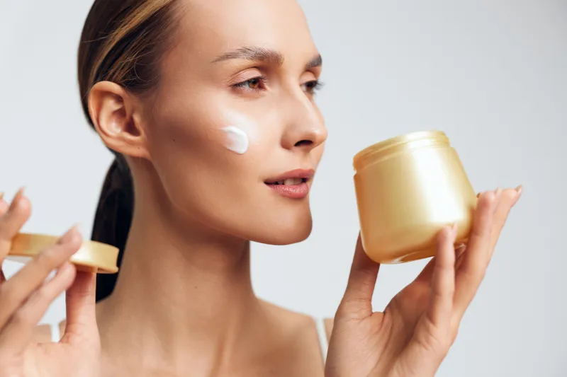 Woman applying face cream to cheek, holding a jar in her hands, close up on light studio background, skincare self care ritual, wellness and beauty mood, calm expression