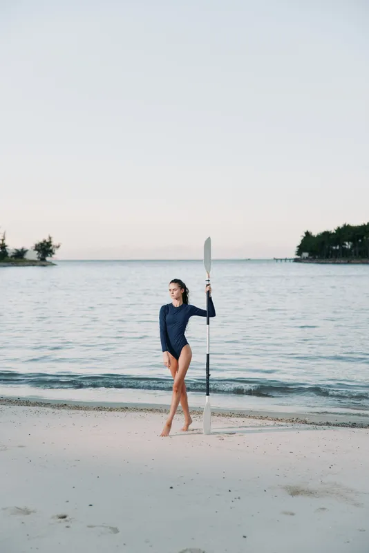 A lone female paddler stands on a serene beach with a paddle, wearing a dark long sleeve swimsuit, as gentle waves lap the shore under a pale, expansive sky.