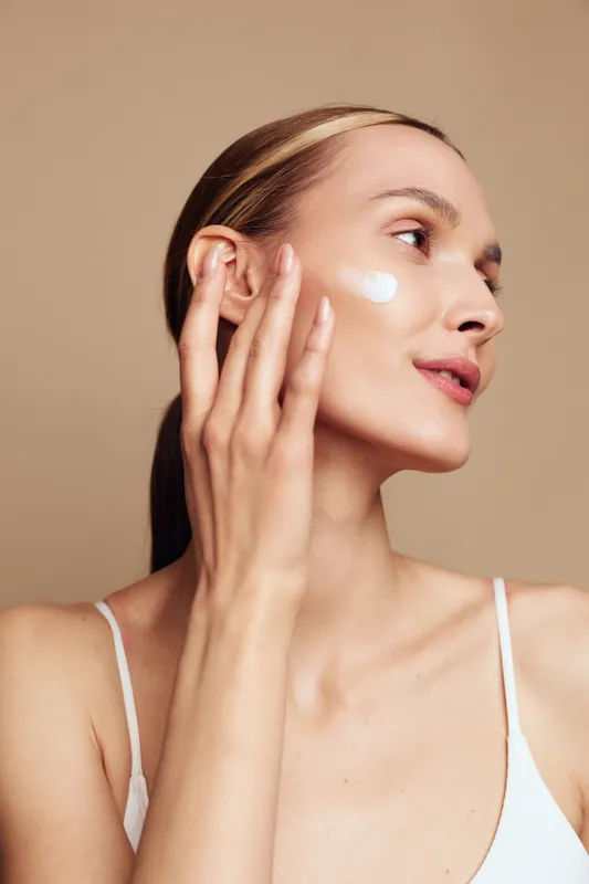 Young woman applying white cream on her cheek, close up portrait against a beige background, skincare routine, self care, gentle massage, smooth skin, soft lighting, calm mood