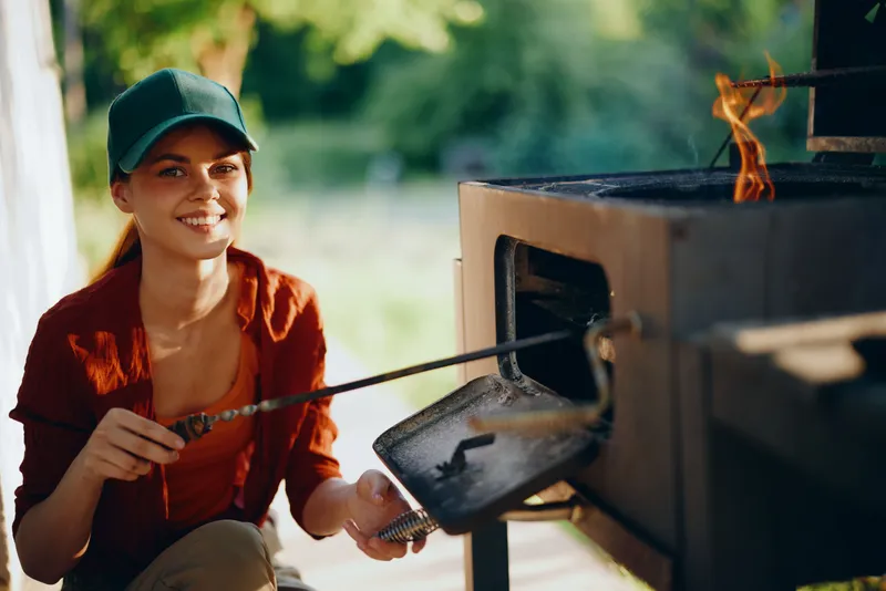 Cheerful young woman in a green cap roasting marshmallows by an outdoor fire pit, enjoying summer vibes and creating memories with friends