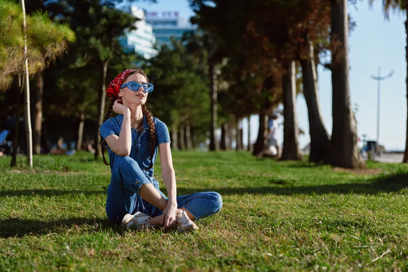 Lifestyle woman in modern boho-western denim street style sits on grass with sunglasses and red bandana, surrounded by trees and sunny outdoor park scene in film color.