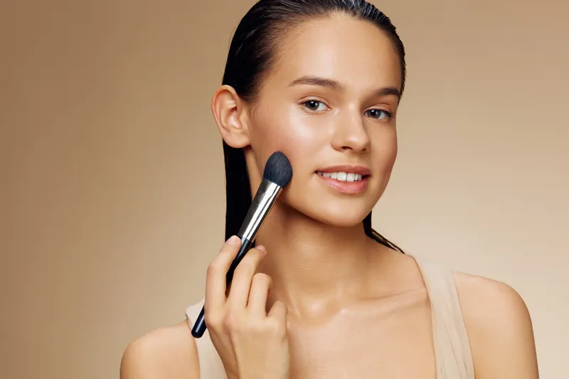 Young model applying makeup with a brush, showcasing natural beauty and confidence against a soft beige background, representing a fresh and radiant look