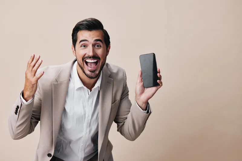Успешный мужчина в деловом костюме держит смартфон, счастливо улыбаясь и делая селфи в студийной обстановке