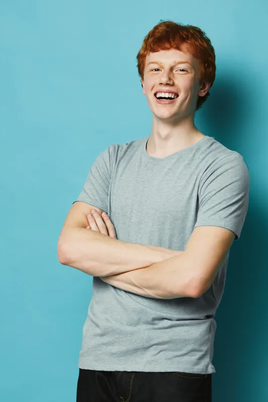 Young man with red hair smiling and standing with crossed arms against a blue background wearing casual grey t shirt and black pants.