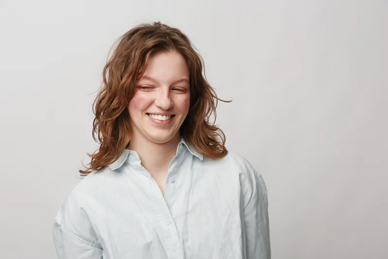 Smiling young woman with curly hair wearing white shirt looks down with closed eyes. Studio portrait on light background. Concept of happiness, natural beauty, and casual lifestyle.