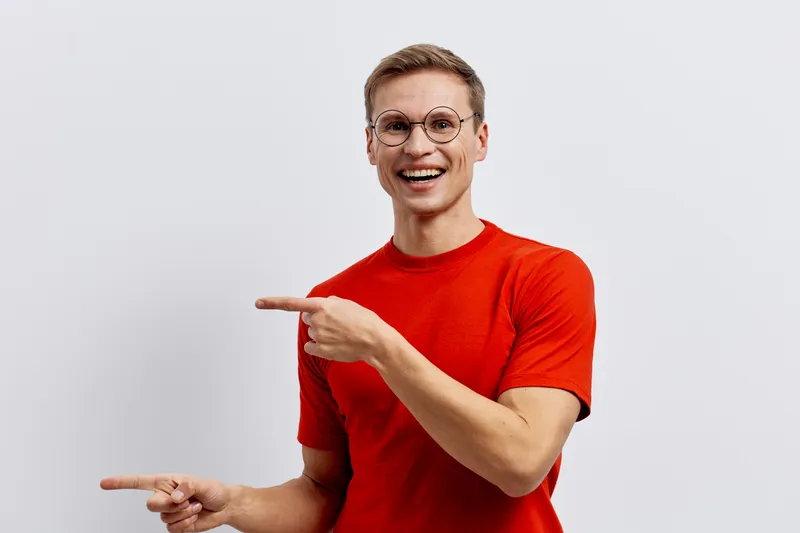 Cheerful young caucasian man with glasses in bright red t-shirt pointing sideways smiling happily isolated on plain white background studio portrait. People lifestyle concept