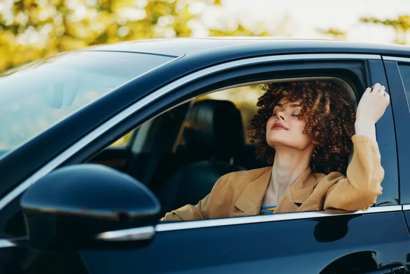 A stylish woman sits in a car with eyes closed, head tilted back, exuding calm and reflection. A portrait of solo travel, relaxed lifestyle, and vintage mood in everyday transportation.