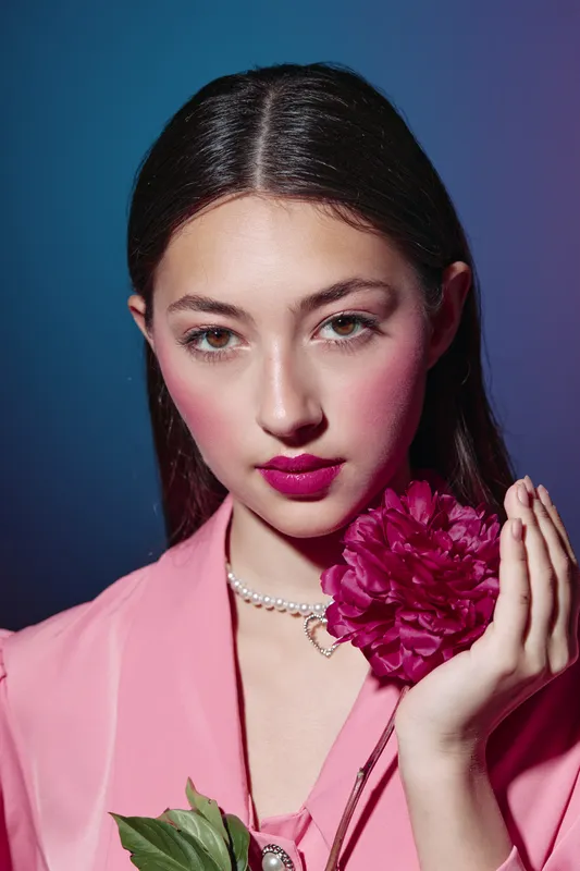 Fashionable young woman with dark hair and bold pink lipstick poses holding deep pink flower close to face against moody dark blue background. Concept of beauty and creative fashion.