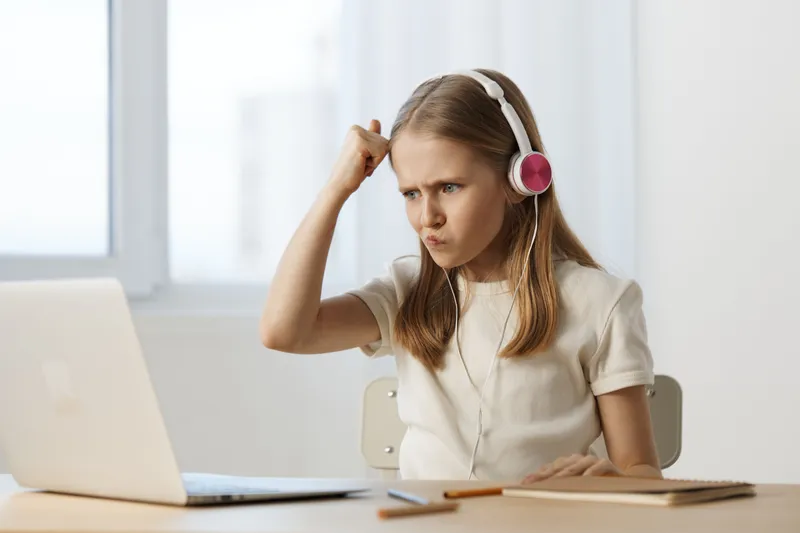 A frustrated young girl with headphones reacts intensely while working on a laptop, showcasing core emotions of concentration and challenge