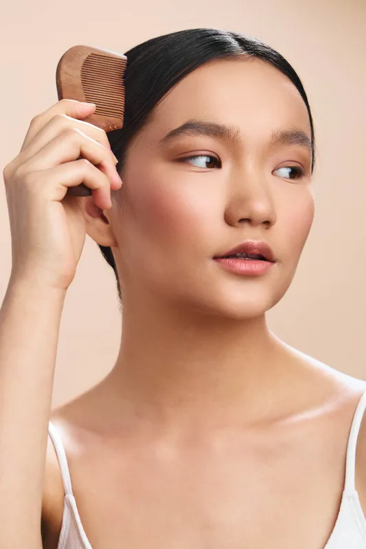 Asian woman brushes her hair in a minimalist studio portrait, soft lighting highlighting smooth skin and calm expression, conveying personal care, empowerment and poised confidence.